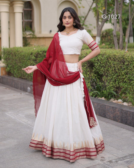 A woman models a traditional Indian outfit consisting of a white crop top, a long white skirt with red and gold borders, and a red dupatta, standing outdoors in front of a building and green hedges. The text 'G-1027' is visible in the top right corner.