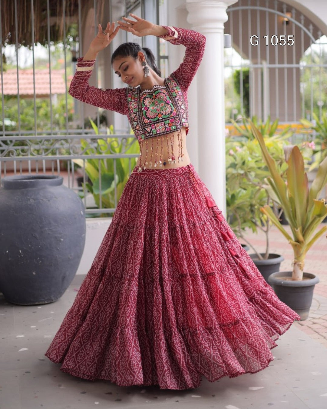 A woman modeling a traditional Indian outfit with a long-sleeved embroidered crop top and a full flared skirt, posed outdoors on a porch with potted plants and columns in the background.
