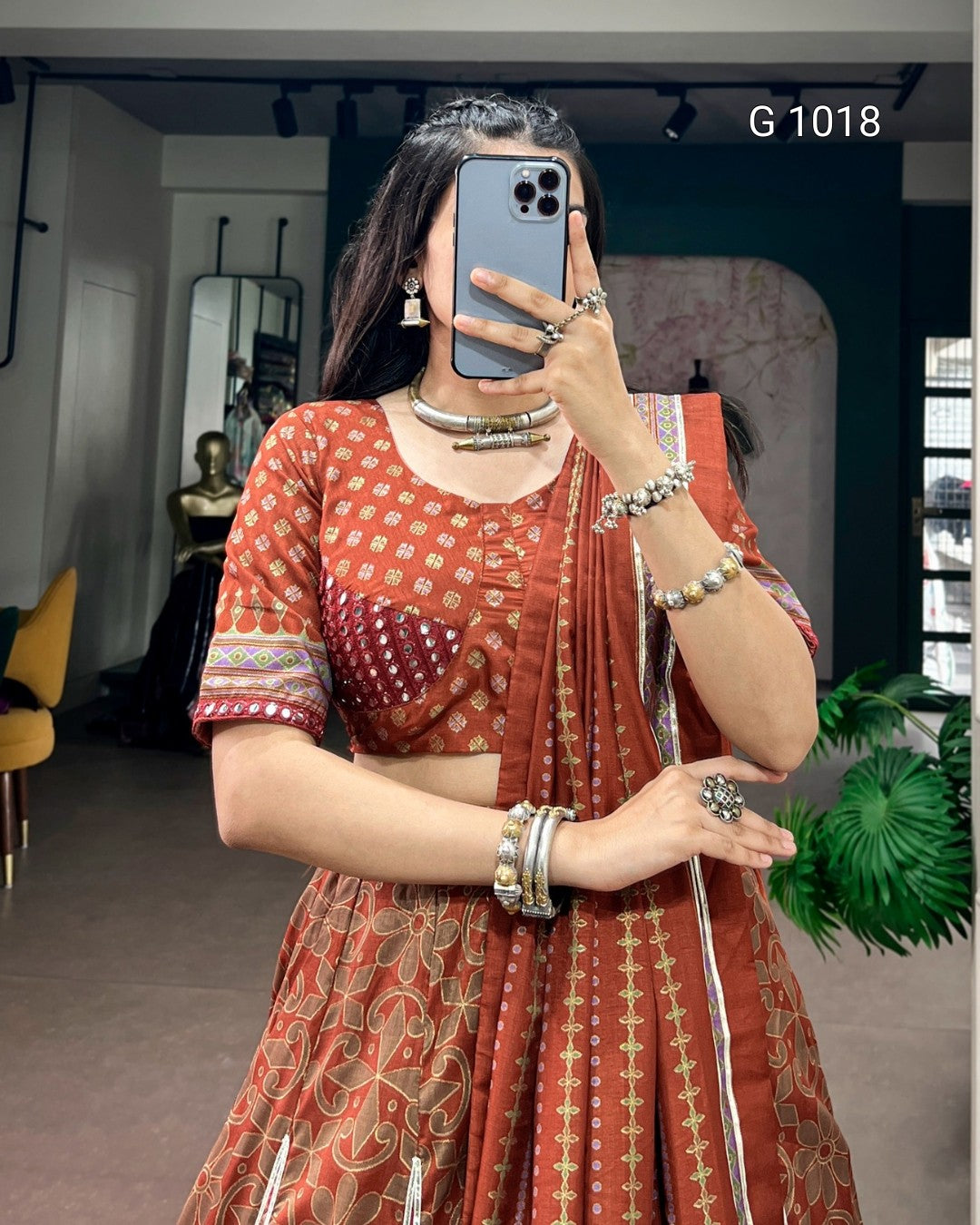 A woman takes a mirror selfie wearing a rust-orange printed traditional Indian outfit with a matching dupatta, silver-toned jewelry, and a blue smartphone. The background shows an indoor boutique setting with mirrors, plants, and furniture.