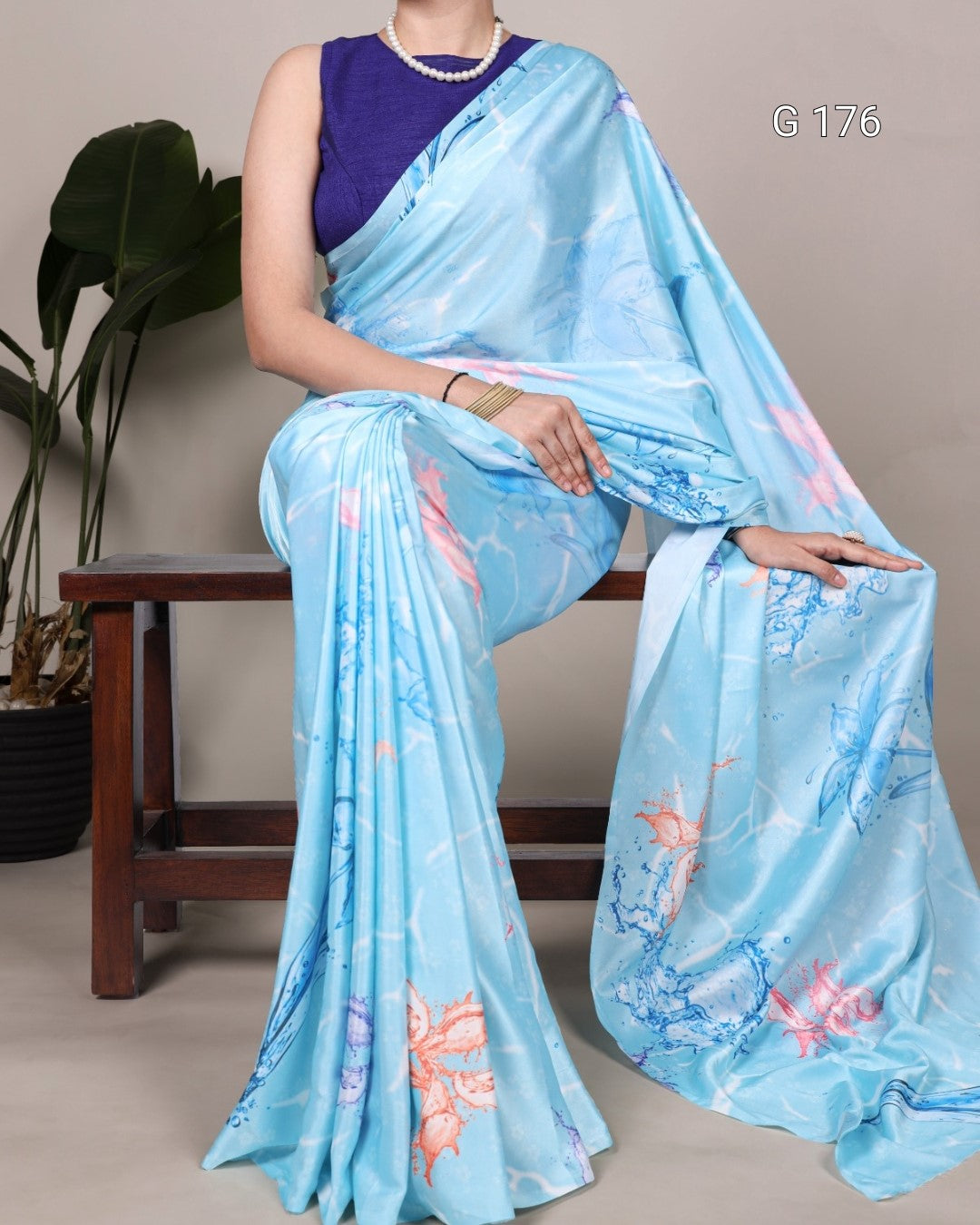 Woman wearing a blue saree with floral patterns, sitting on a wooden stool against a plain background.