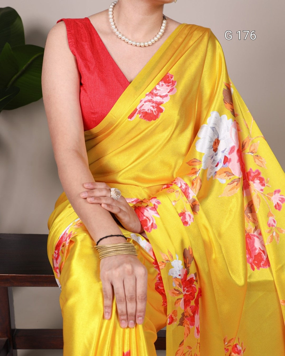 Woman wearing a yellow saree with floral patterns and a red blouse, sitting against a neutral background.