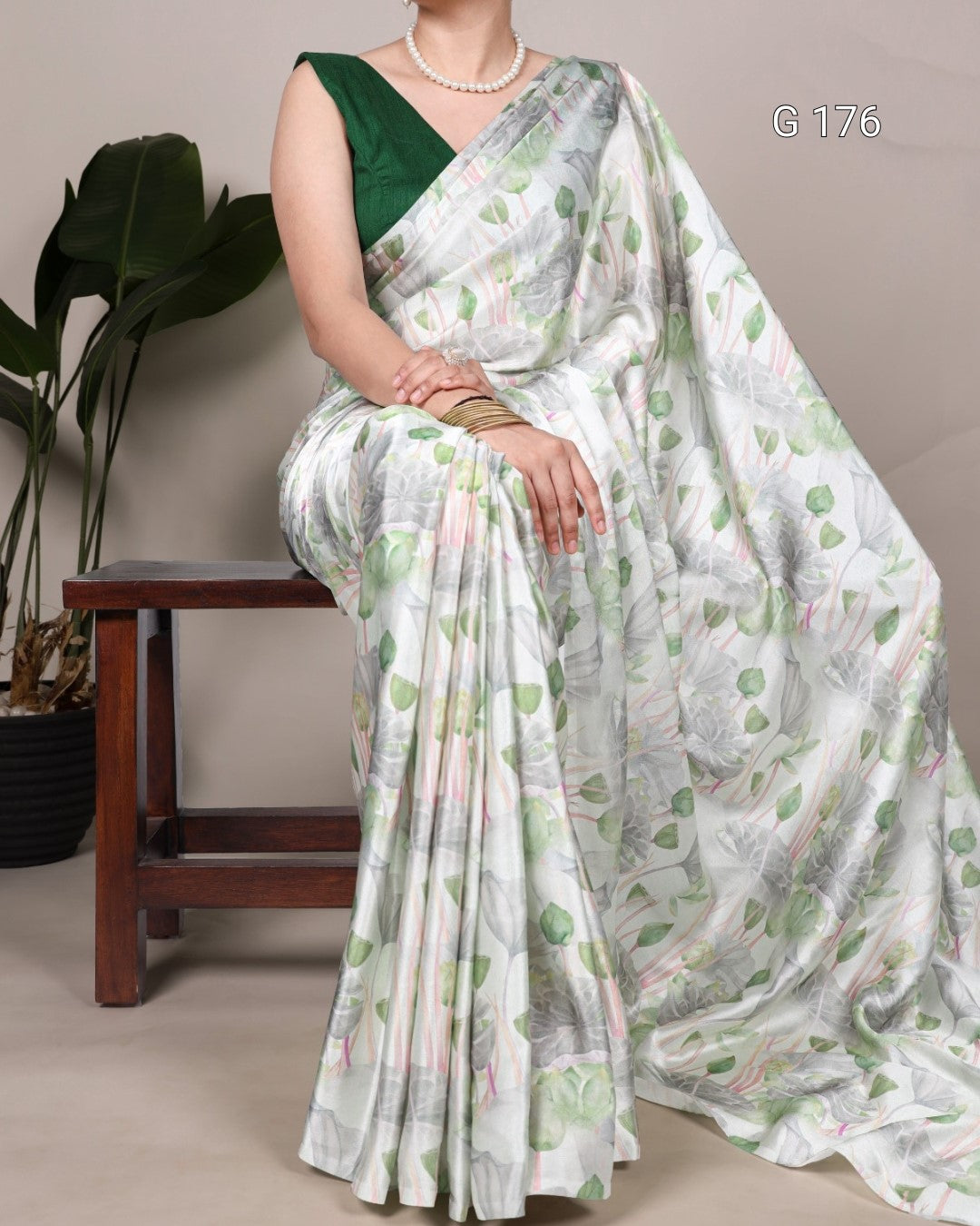 Woman wearing a white saree with green leaf patterns, sitting on a wooden stool.