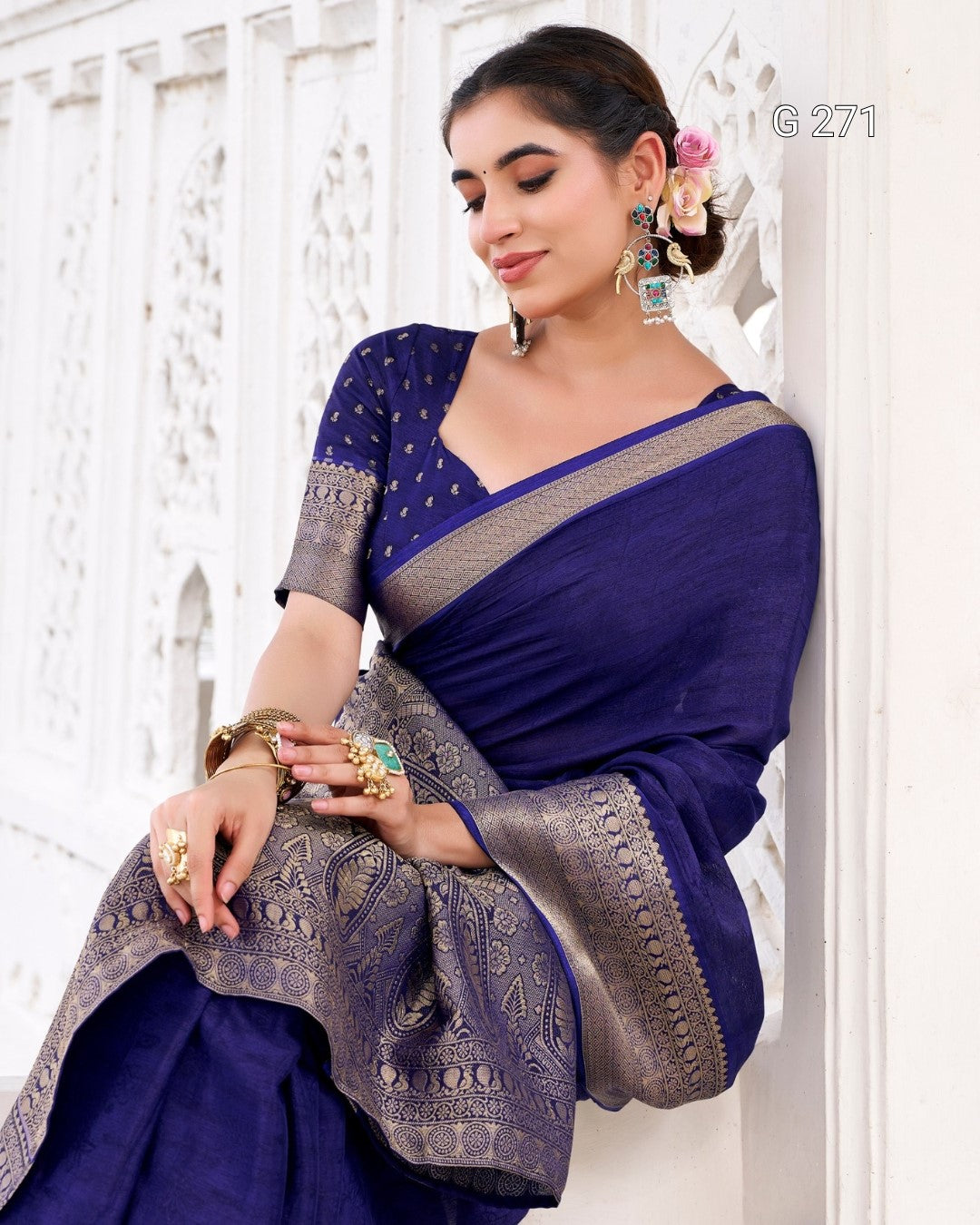 A woman wearing a royal blue saree with a silver patterned border and matching embroidered blouse, posed against a white carved wall.