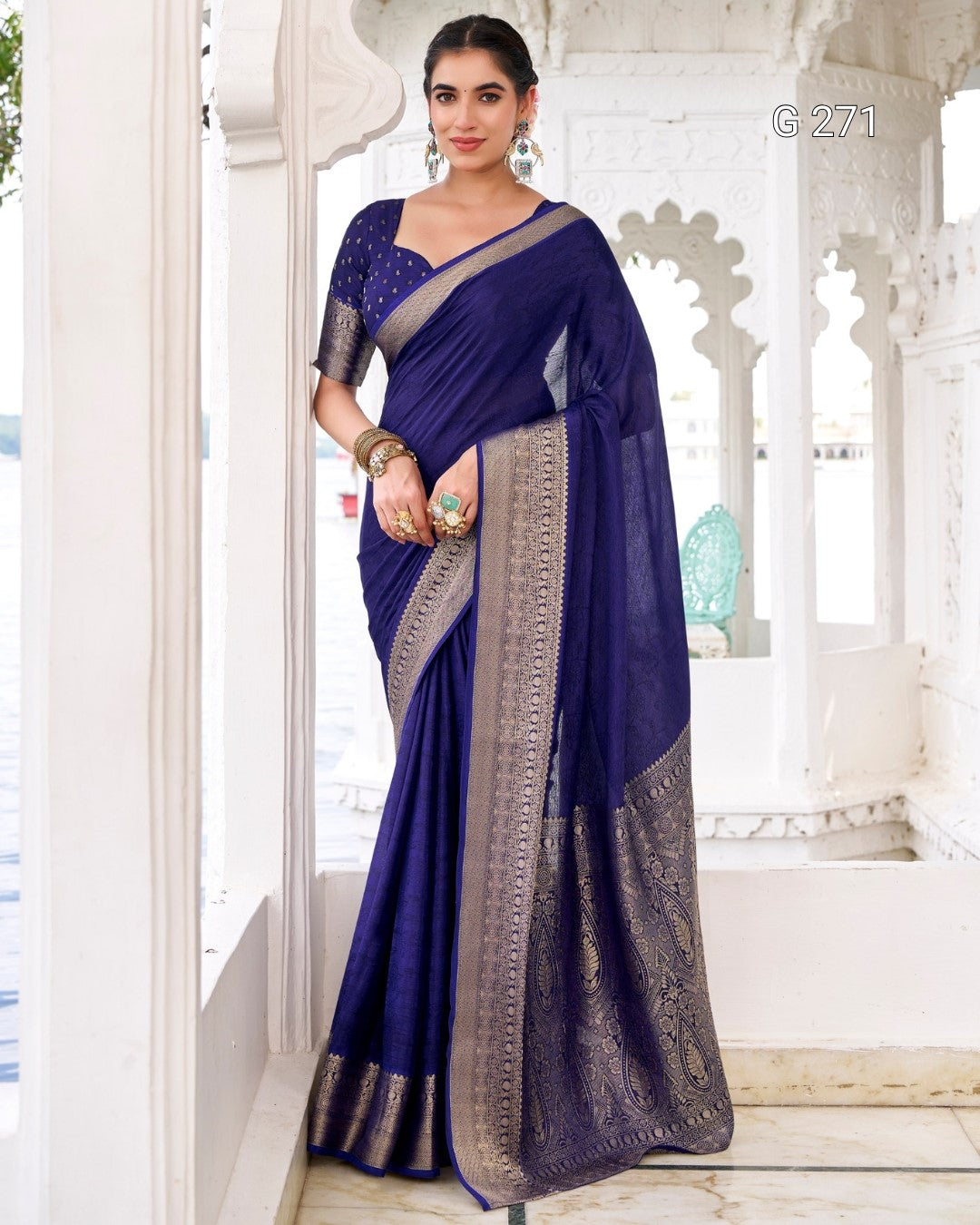 A woman models a royal blue silk saree with a wide metallic silver border and a matching blue embroidered blouse, standing in a white architectural corridor.