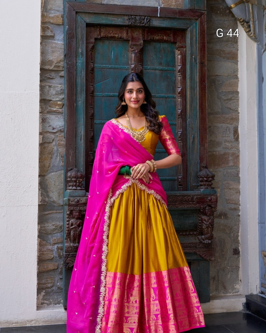 Woman in a pink and yellow traditional outfit standing in front of a stone wall.