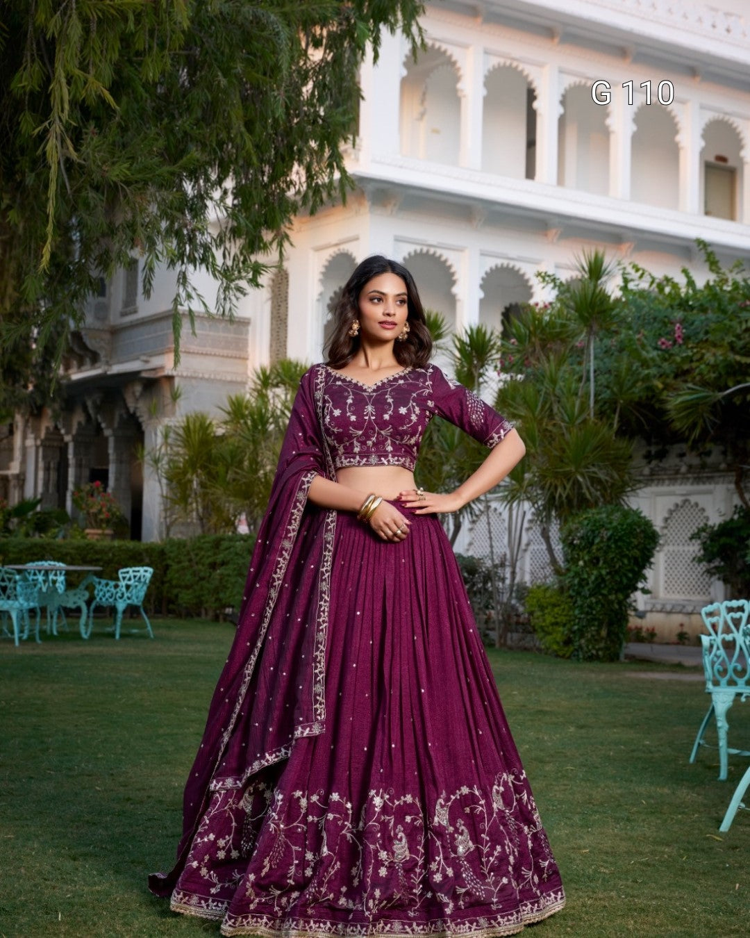 Woman in a purple traditional outfit standing in front of a building with greenery.