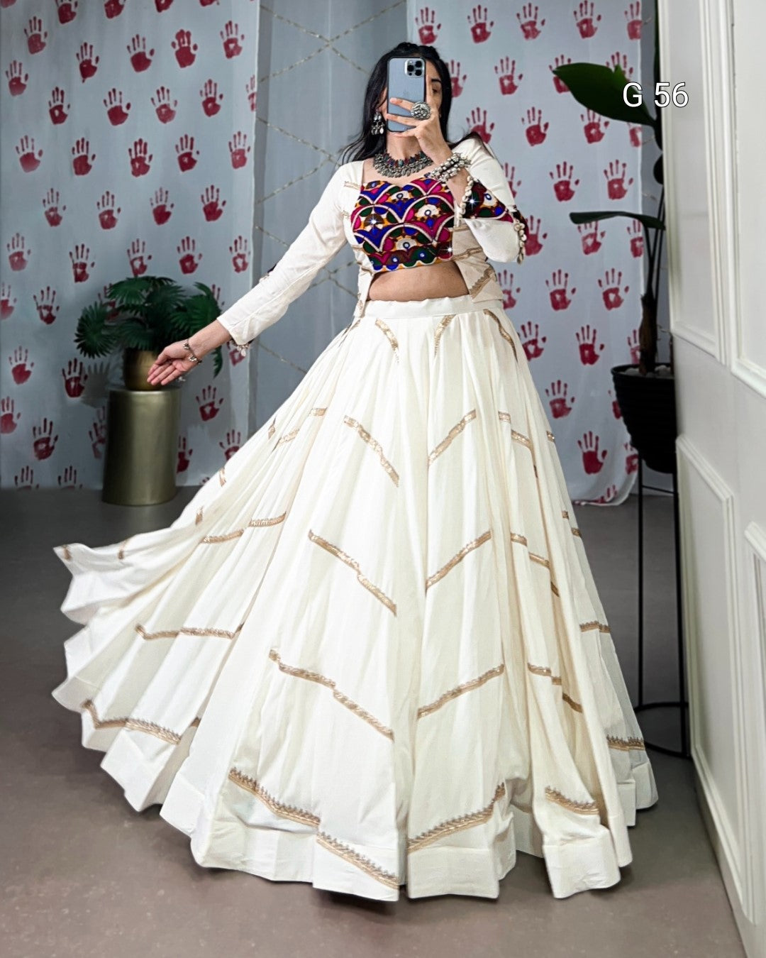 Person wearing a traditional outfit with a white skirt and multicolored top in a room with a patterned wall.