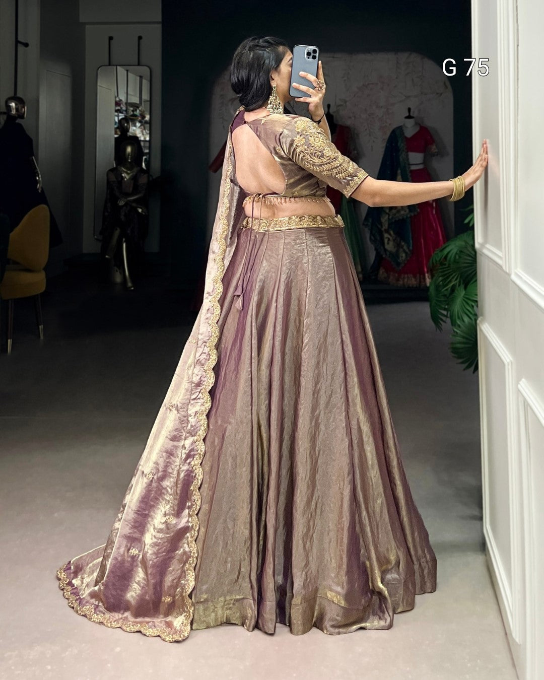Woman in a traditional outfit taking a mirror selfie in a room with other people and clothing in the background.