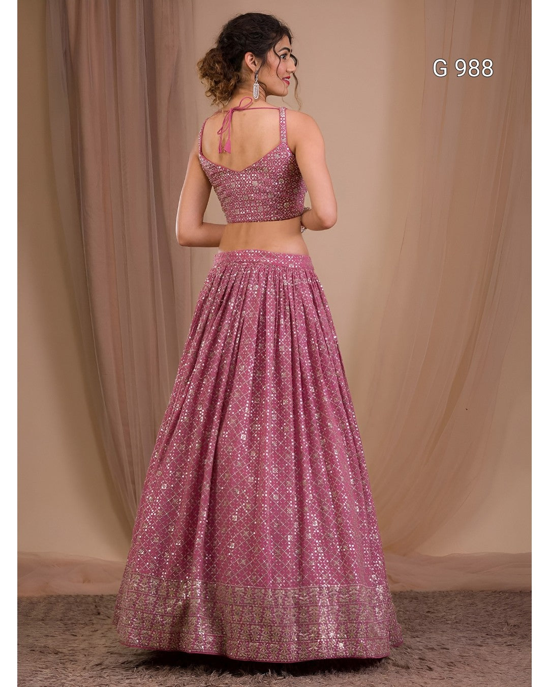 A woman wearing a pink heavily embellished lehenga choli set, photographed from the back against a draped beige fabric backdrop.