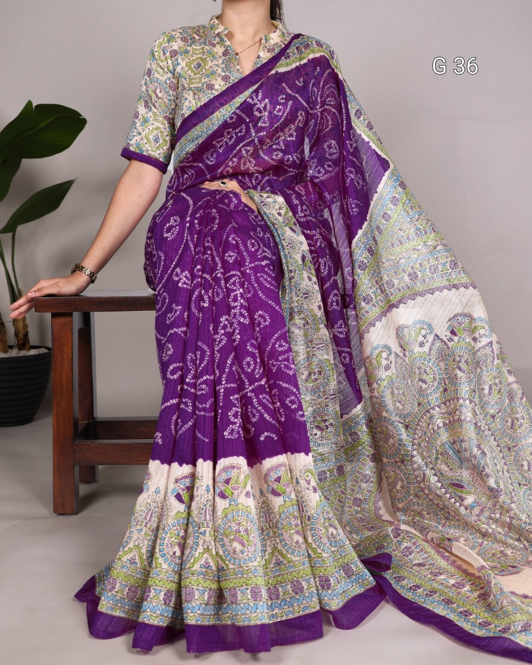 Woman wearing a purple and white patterned saree with a matching blouse, sitting on a wooden stool.