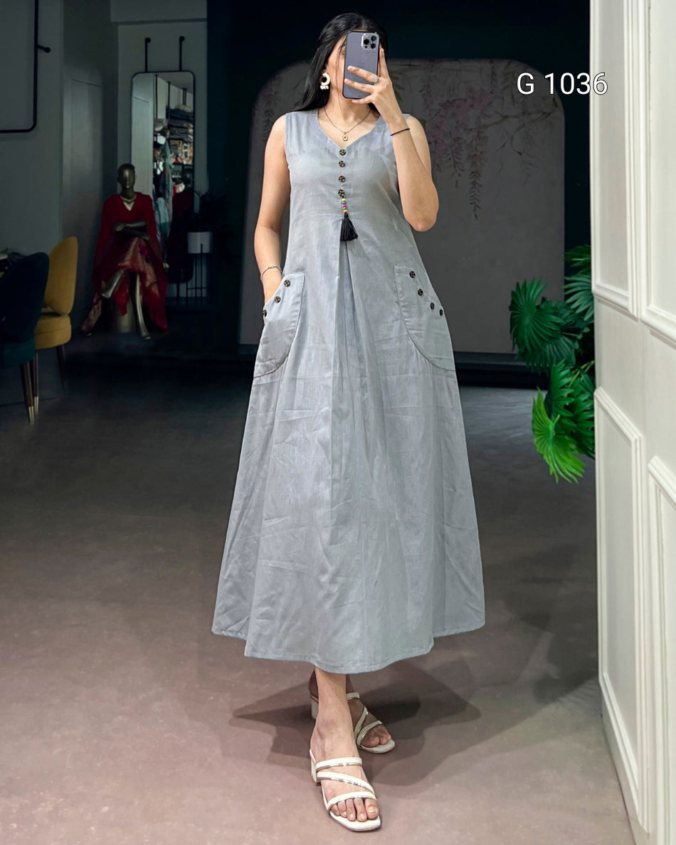 A woman takes a mirror selfie wearing a sleeveless, light gray linen maxi dress with a button-down front and large patch pockets. The setting is a boutique interior with a gray floor, a mannequin in the background, and a white door frame on the right.