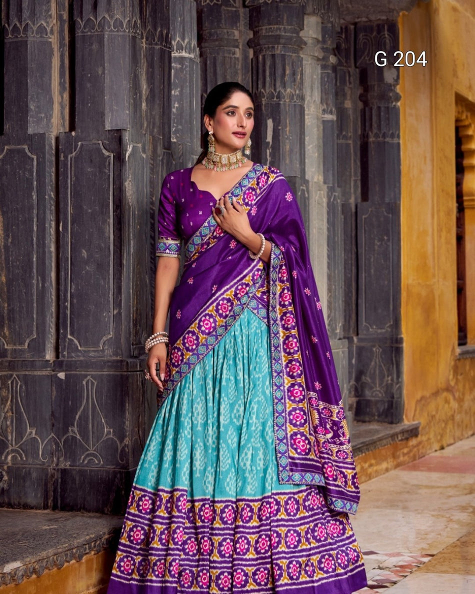 Woman wearing a traditional purple and teal saree with a decorative pattern, standing in front of a dark stone wall.
