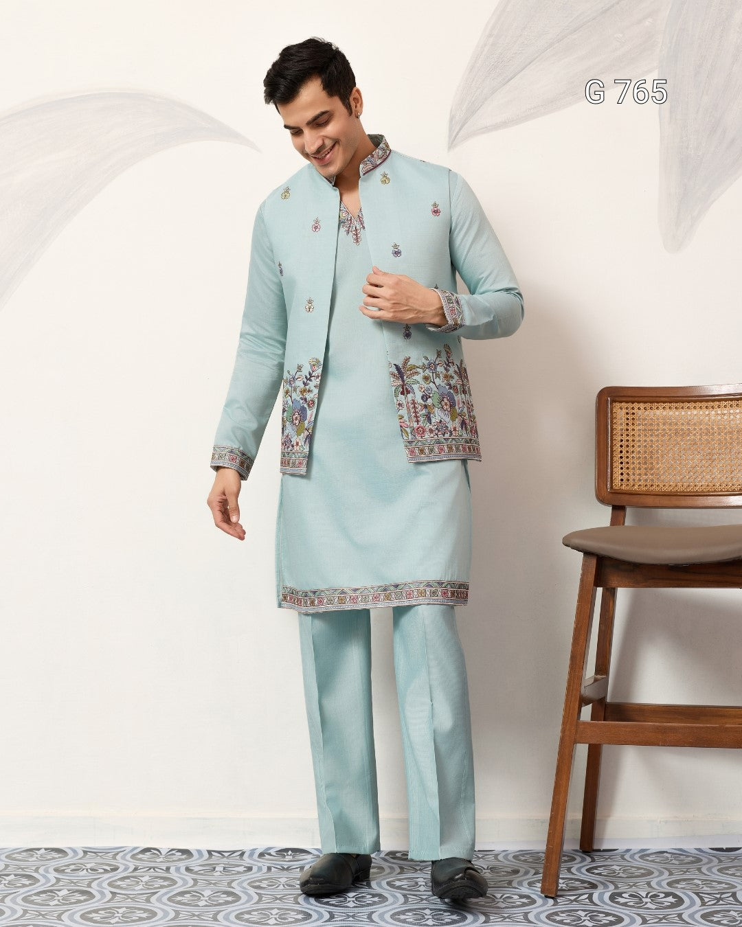 Man wearing a light blue traditional outfit with intricate patterns, standing against a white wall.