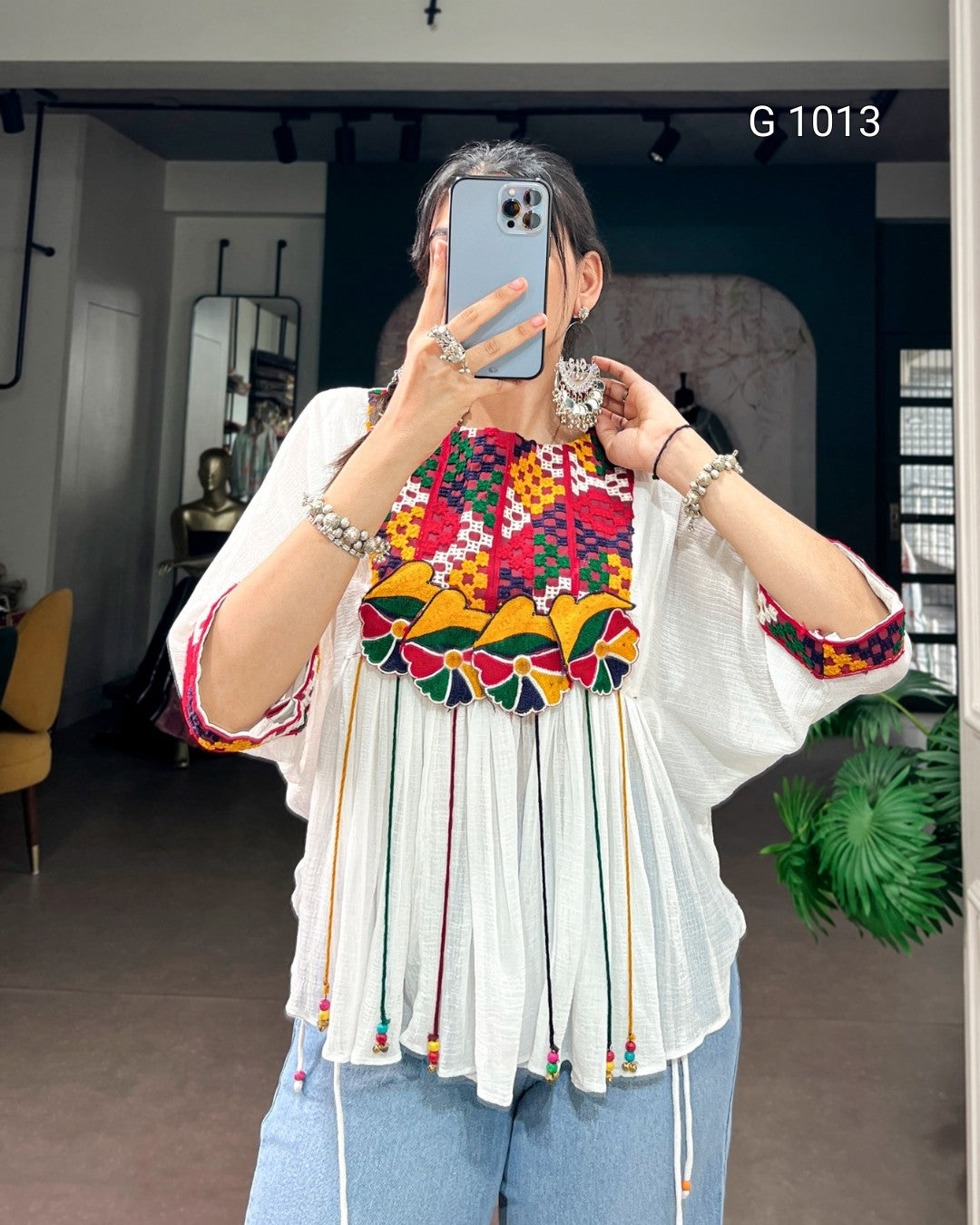 A woman takes a mirror selfie in a boutique wearing a white blouse with a vibrant, multicolored geometric embroidered yoke and colorful fringe details. She is wearing blue jeans and holding a blue smartphone.