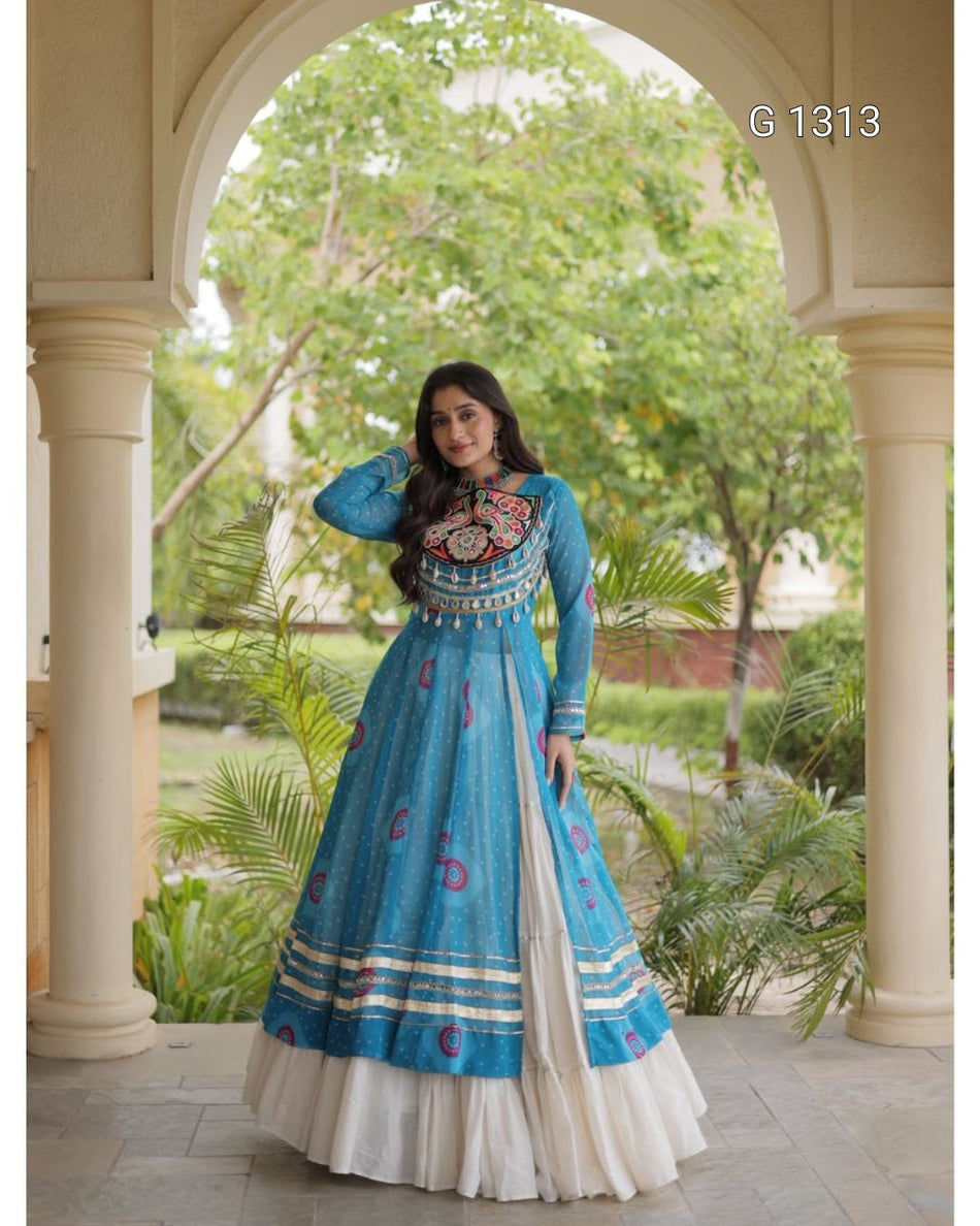 A woman wearing a blue and white traditional Anarkali gown stands under a stone archway with green garden foliage in the background. The dress features intricate embroidery on the bodice and border details, with a white pleated underskirt visible at the hem.