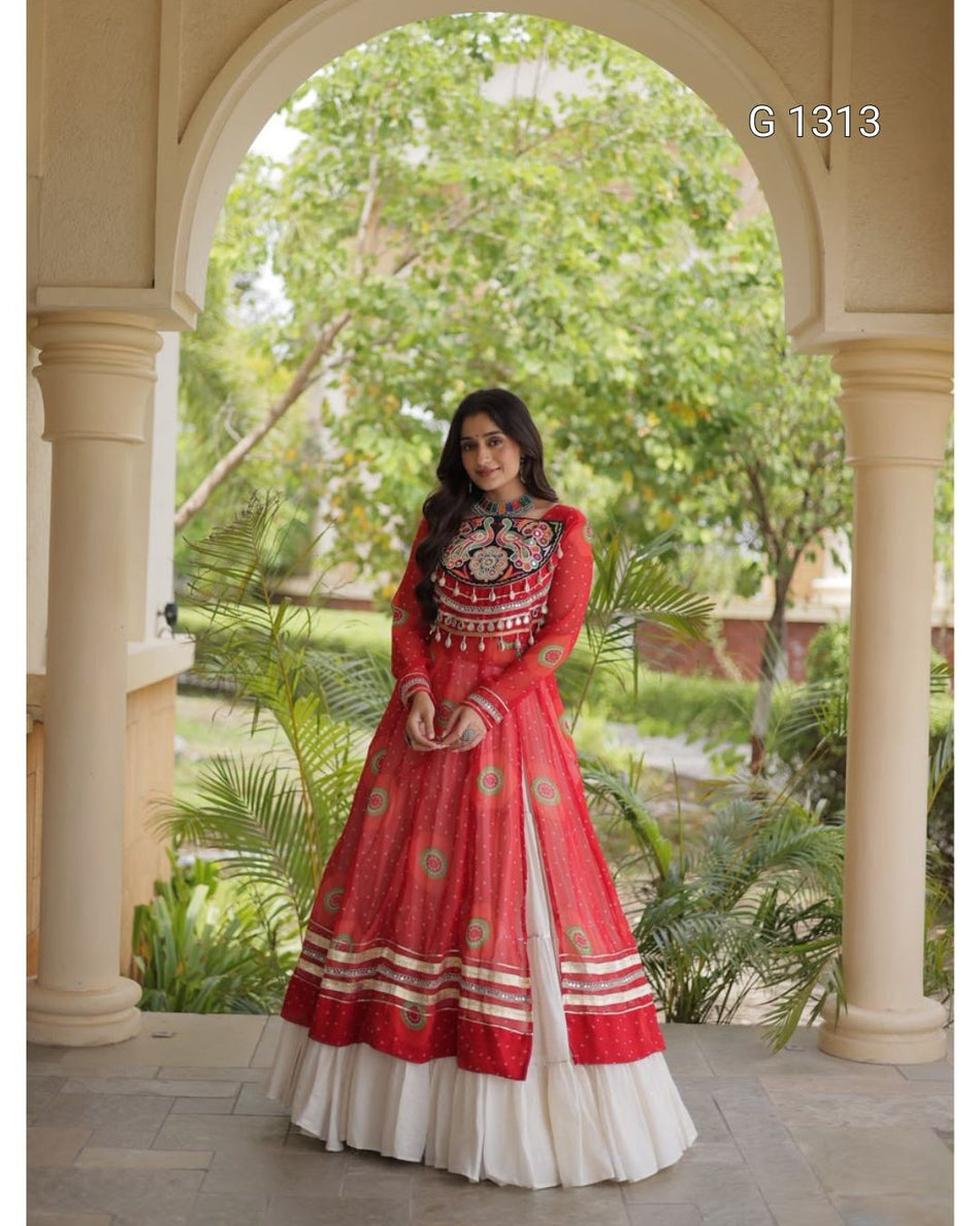 A woman wearing a red and white embroidered Anarkali-style dress stands under a large stone archway with green garden foliage in the background. The dress features a red upper bodice with intricate multicolor embroidery, a red flared skirt with gold and red patterns, and a layered white underskirt.