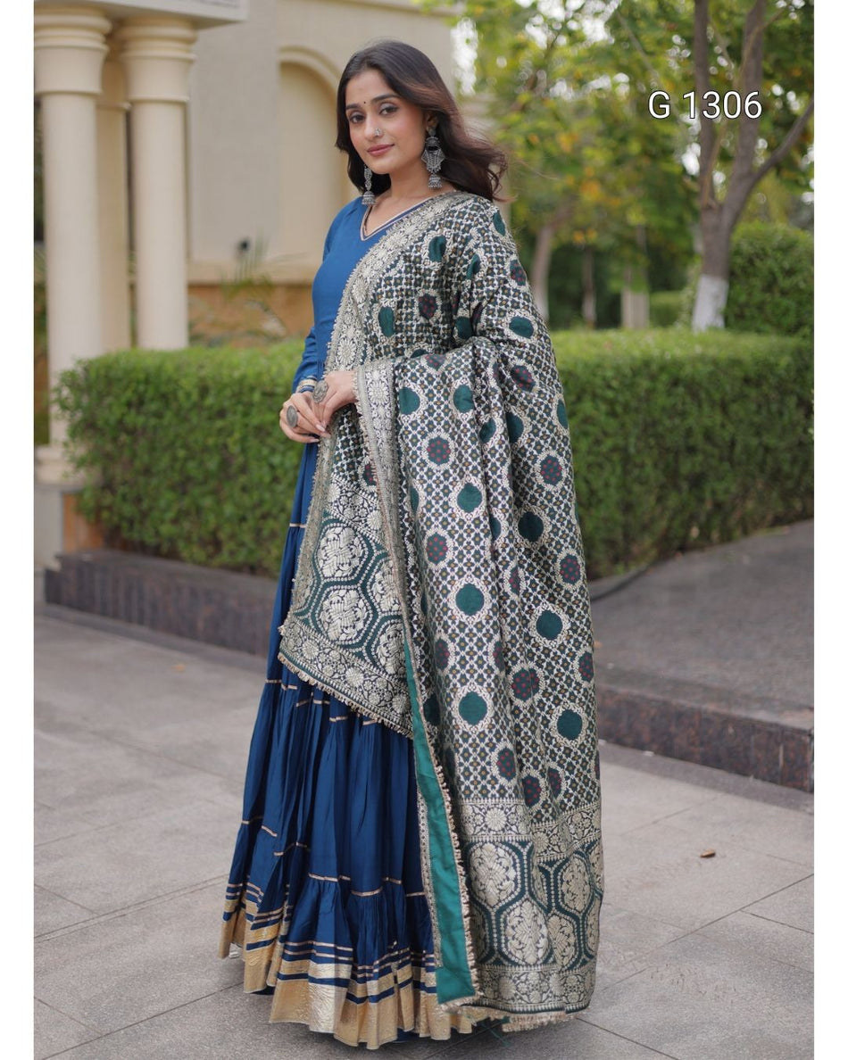 A woman models a traditional Indian outfit consisting of a navy blue pleated skirt (lehenga), a solid blue blouse, and a heavily embroidered gray dupatta with green and gold geometric patterns. The background shows a paved walkway, green hedges, and a building with columns.