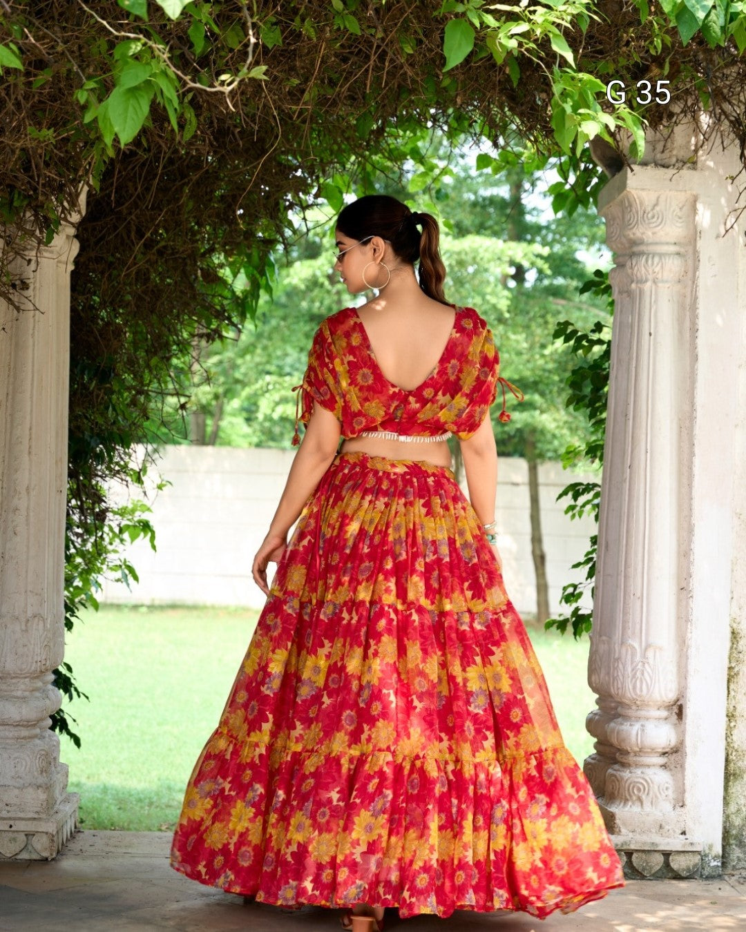Woman in a red and yellow patterned dress standing under a stone archway with greenery.