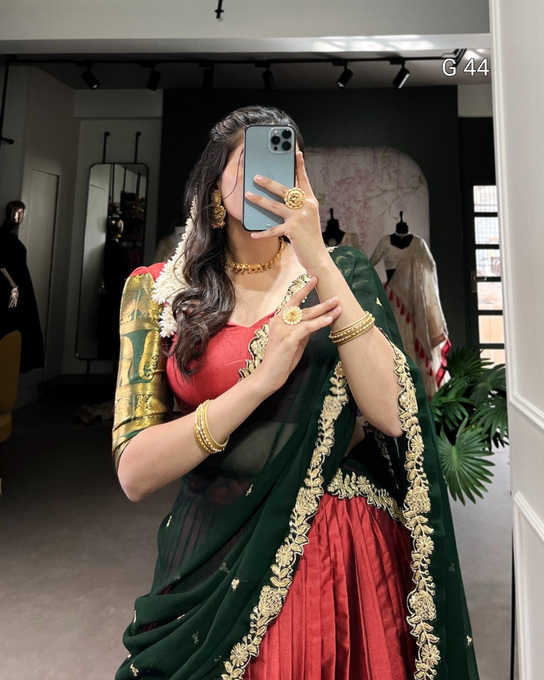 Woman taking a mirror selfie wearing a green and red saree with gold jewelry.