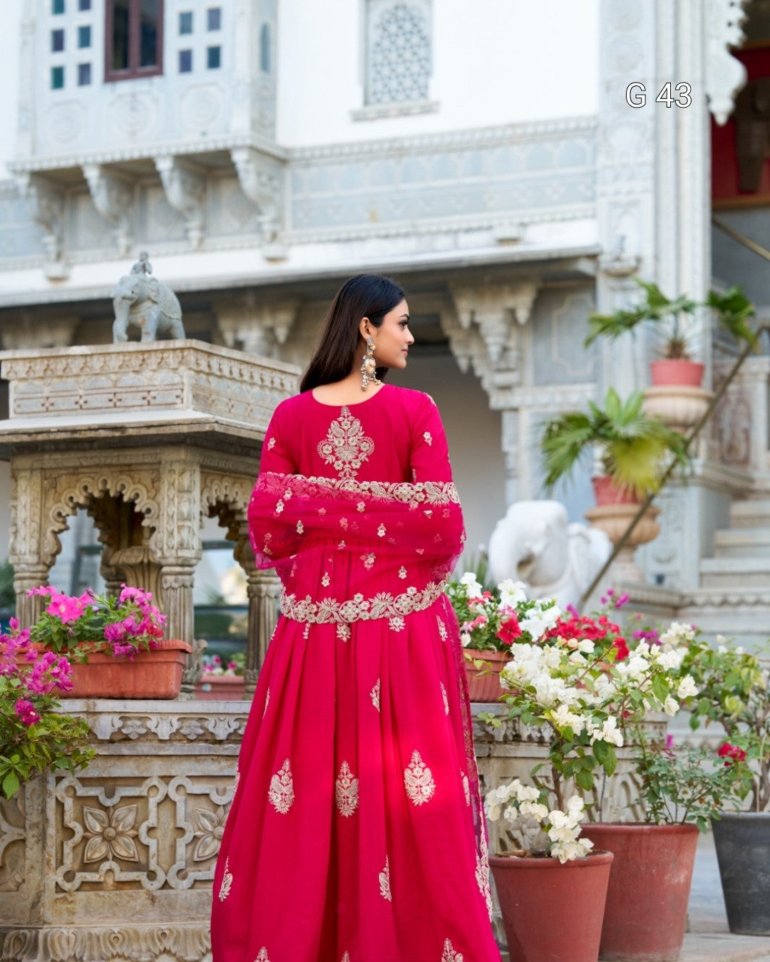 Woman in a red traditional outfit standing in front of an architectural building with flowers around.