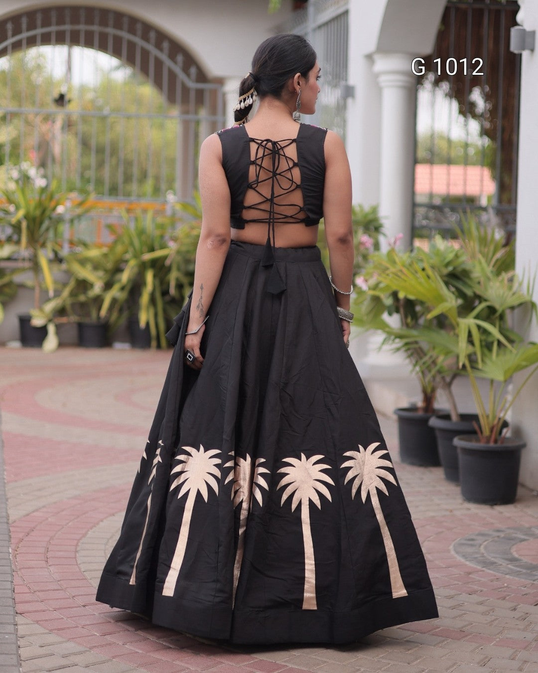 A woman viewed from behind wearing a black sleeveless maxi gown with a lace-up open back and a flared skirt featuring large beige palm tree motifs near the hem, photographed outdoors in front of a building with potted plants.