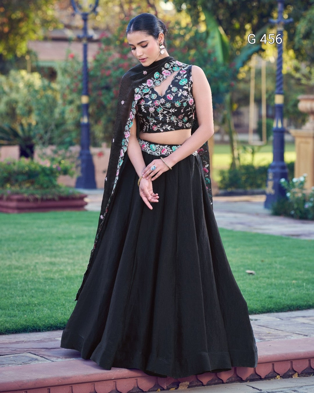 A woman stands outdoors in a garden wearing a black traditional outfit with floral embroidery on the bodice and a matching draped scarf. The setting includes green grass, trees, and a paved walkway.