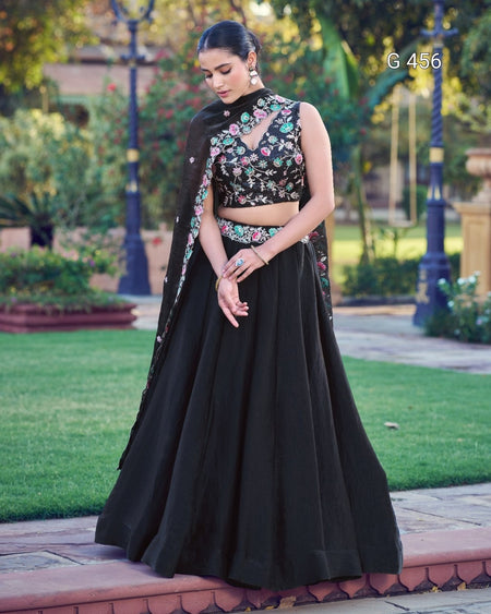 A woman stands outdoors in a garden wearing a black traditional outfit with floral embroidery on the bodice and a matching draped scarf. The setting includes green grass, trees, and a paved walkway.