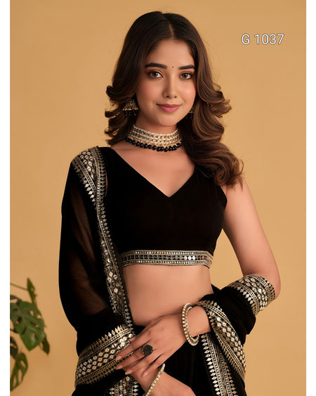 A woman modeling a black traditional outfit with ornate silver mirror-work embroidery on the blouse border, dupatta, and wristbands, posed against a solid beige background.