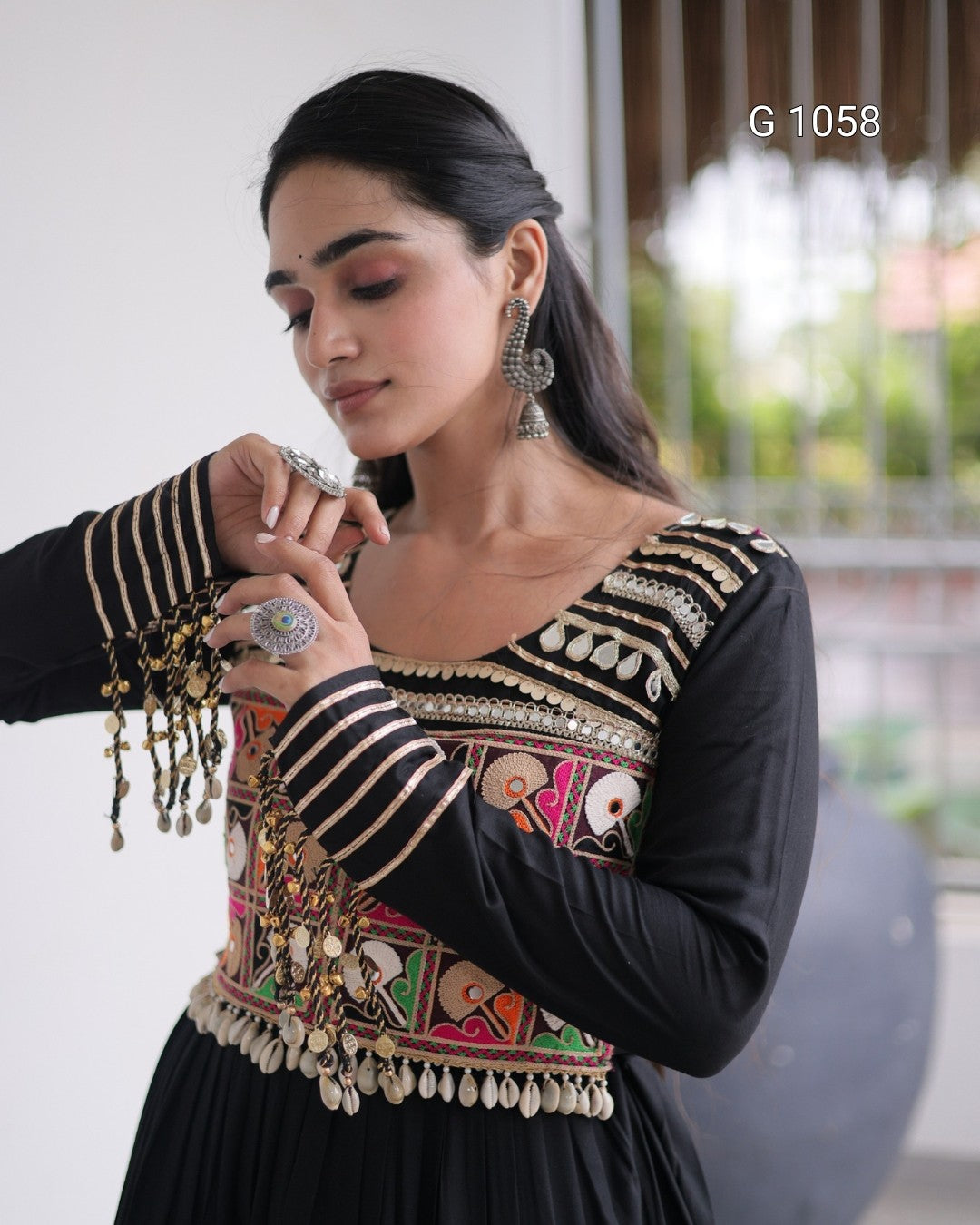A woman wearing a black long-sleeve embroidered dress with intricate multicolor floral and geometric patterns, silver coin embellishments, and striped cuffs. She is shown looking down while adjusting her sleeve, with a soft-focus indoor background.