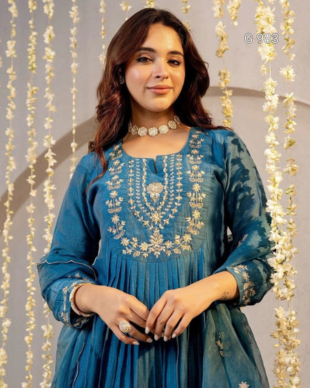 A woman wearing a teal blue traditional embroidered dress with intricate gold embroidery on the chest, standing in front of a decorative backdrop of hanging white flower garlands.