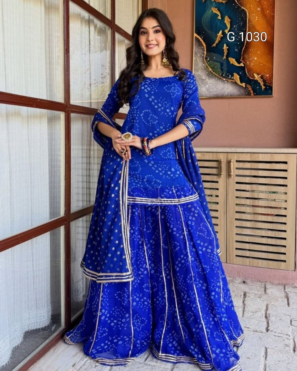 A woman models a royal blue traditional Indian outfit with white printed motifs and silver borders, consisting of a kurta, flared pants, and a dupatta. She is standing in an indoor setting with a window and wall art in the background.