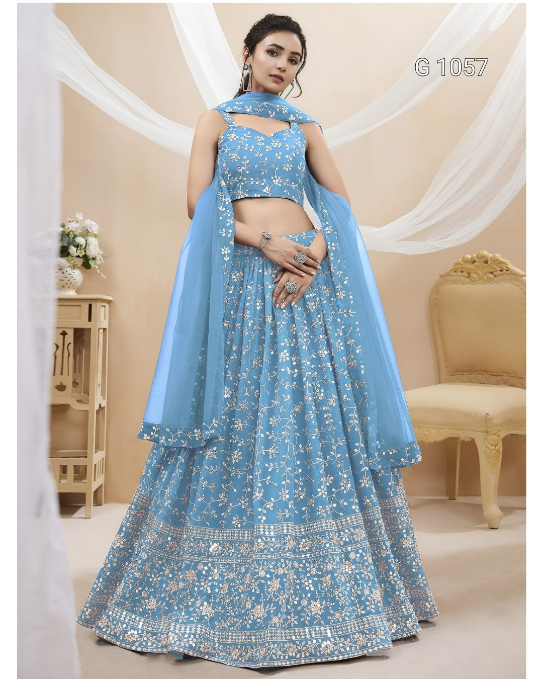 A woman models a light blue embroidered Indian ethnic outfit with a sleeveless crop top, flared skirt, and matching dupatta in a decorated indoor setting.
