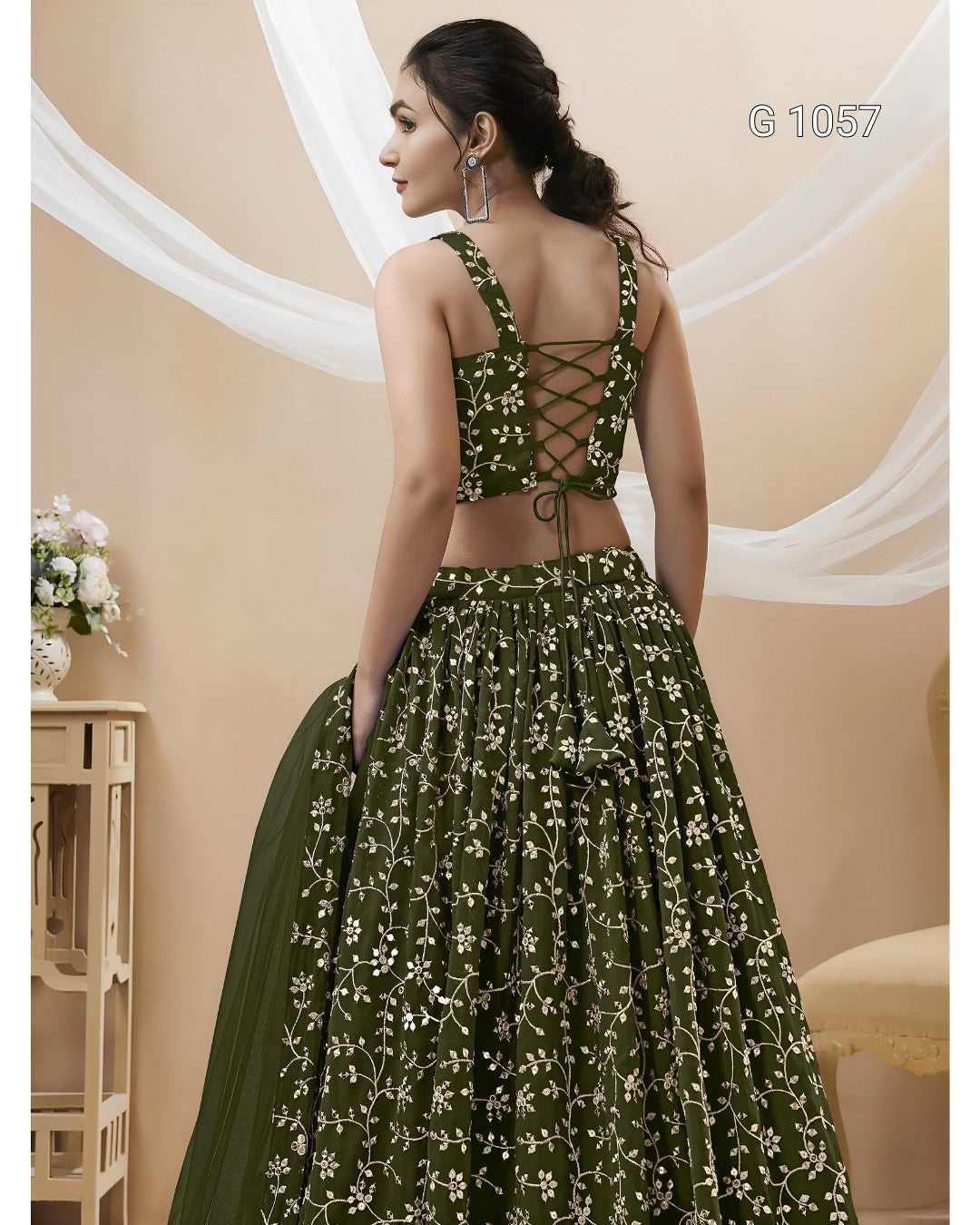 A woman modeling a dark olive green lehenga choli with white floral embroidery, shown from the back. The dress features a lace-up crop top and a full skirt, with a matching dupatta draped over one side.