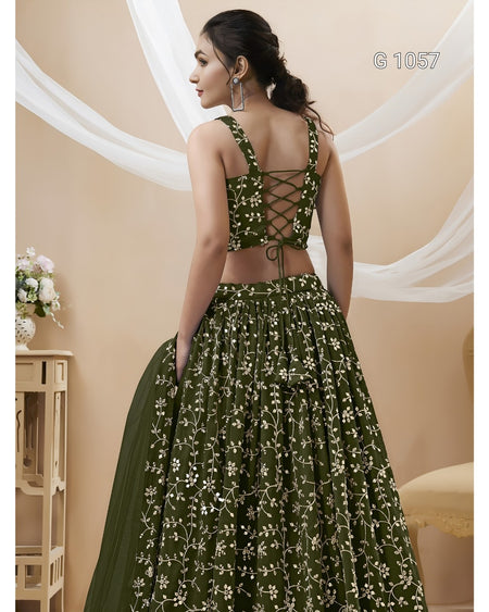 A woman modeling a dark olive green lehenga choli with white floral embroidery, shown from the back. The dress features a lace-up crop top and a full skirt, with a matching dupatta draped over one side.