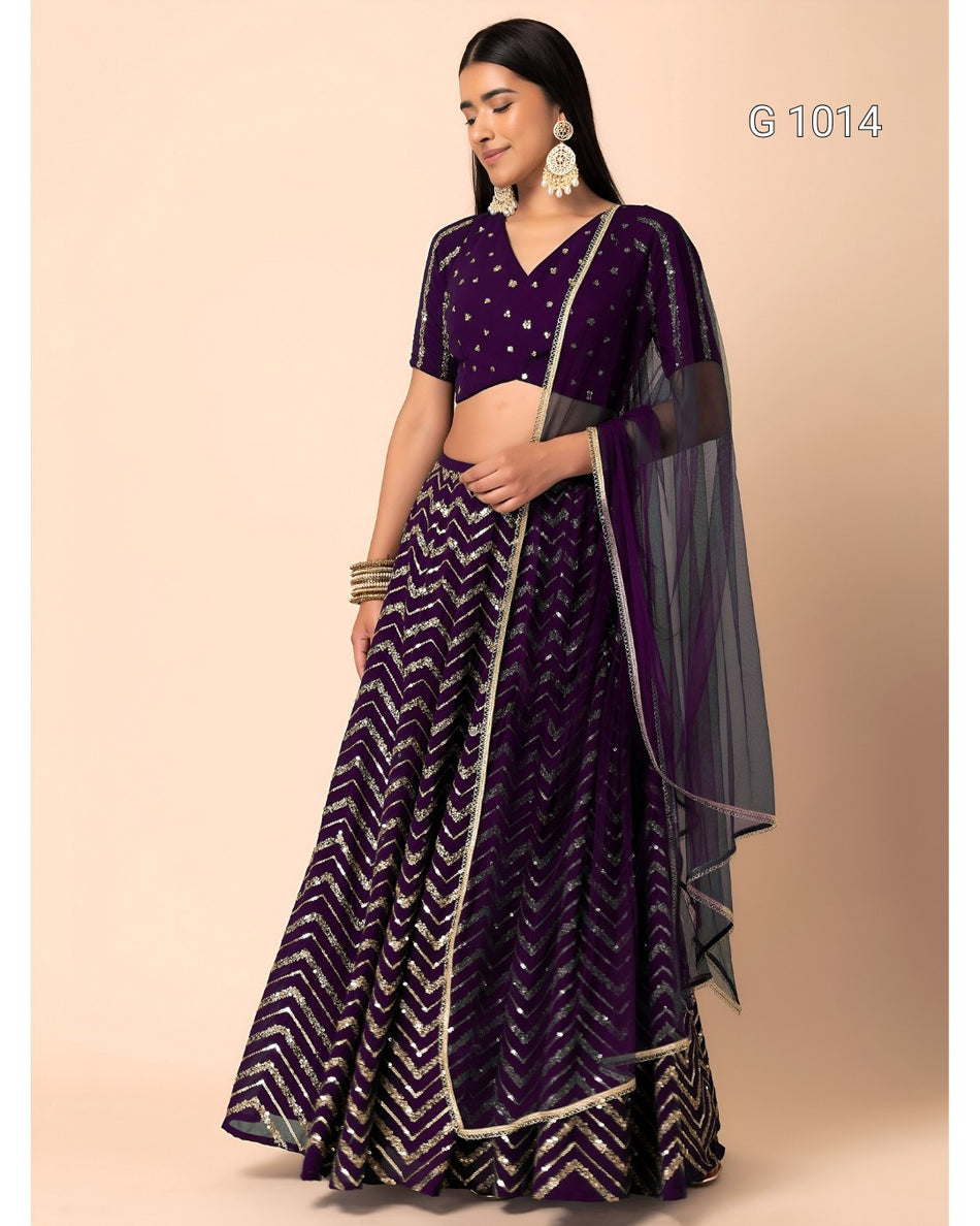 A woman modeling a traditional Indian ethnic outfit consisting of a dark purple cropped blouse with silver embellishments, a matching flared skirt with a silver chevron pattern, and a sheer purple dupatta, set against a solid beige background.