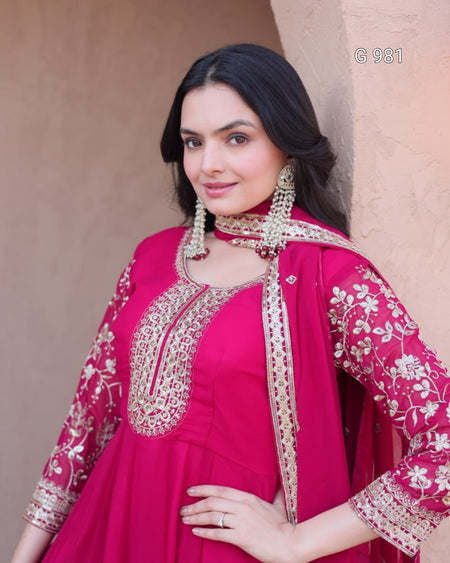A woman wearing a bright pink traditional outfit with ornate gold embroidery and a matching embroidered dupatta, posed against a beige stucco wall.
