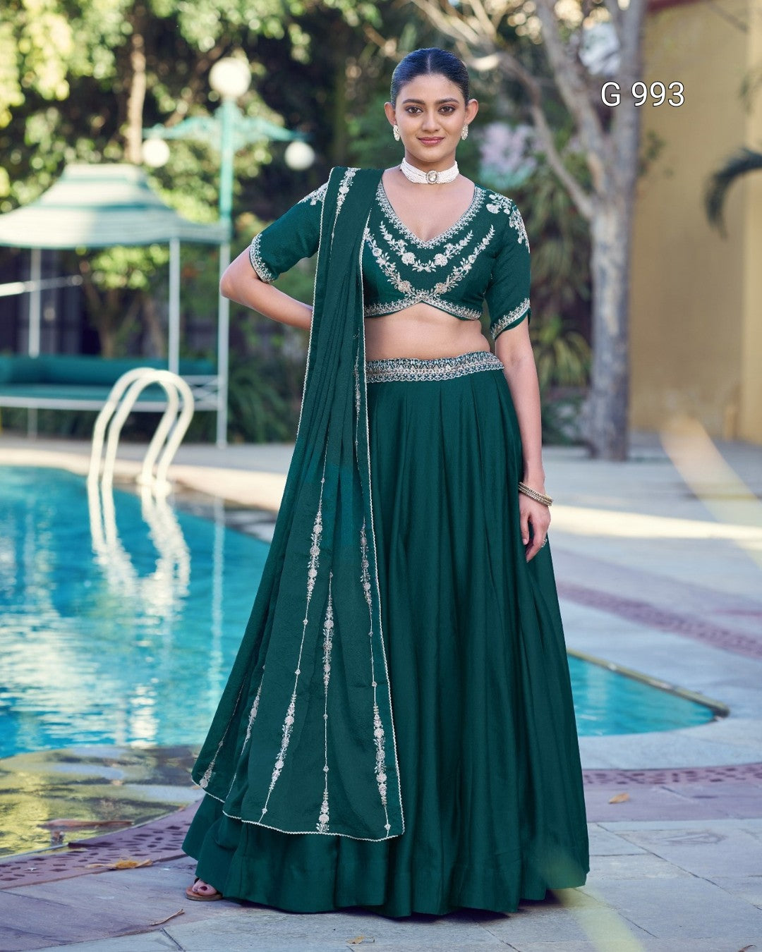 A woman stands outdoors beside a swimming pool wearing a dark green traditional outfit with a matching draped scarf, embellished blouse, and flared skirt. The background includes pool water, trees, and patio seating.