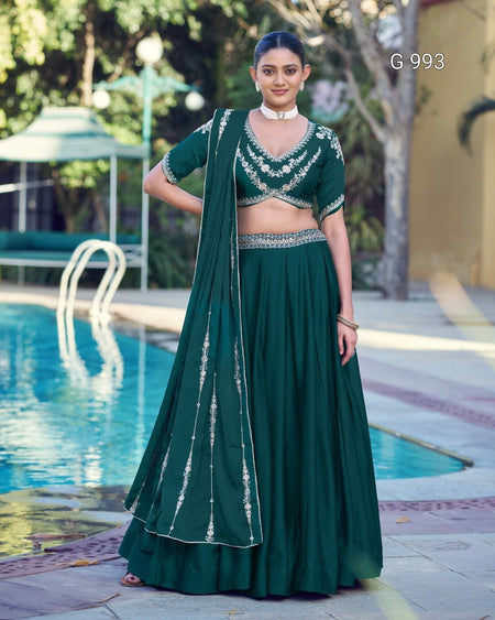 A woman stands outdoors beside a swimming pool wearing a dark green traditional outfit with a matching draped scarf, embellished blouse, and flared skirt. The background includes pool water, trees, and patio seating.