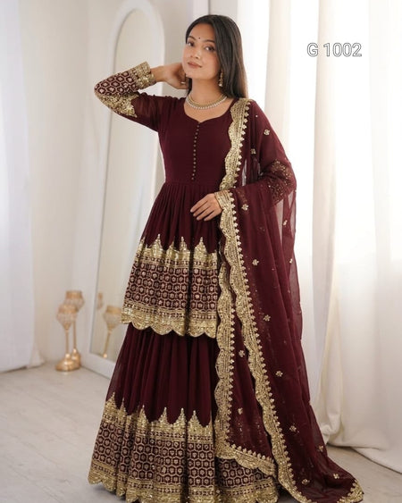A woman models a deep maroon Anarkali gown with intricate gold embroidery and a matching dupatta, set against a soft, cream-colored indoor background with a mirror.