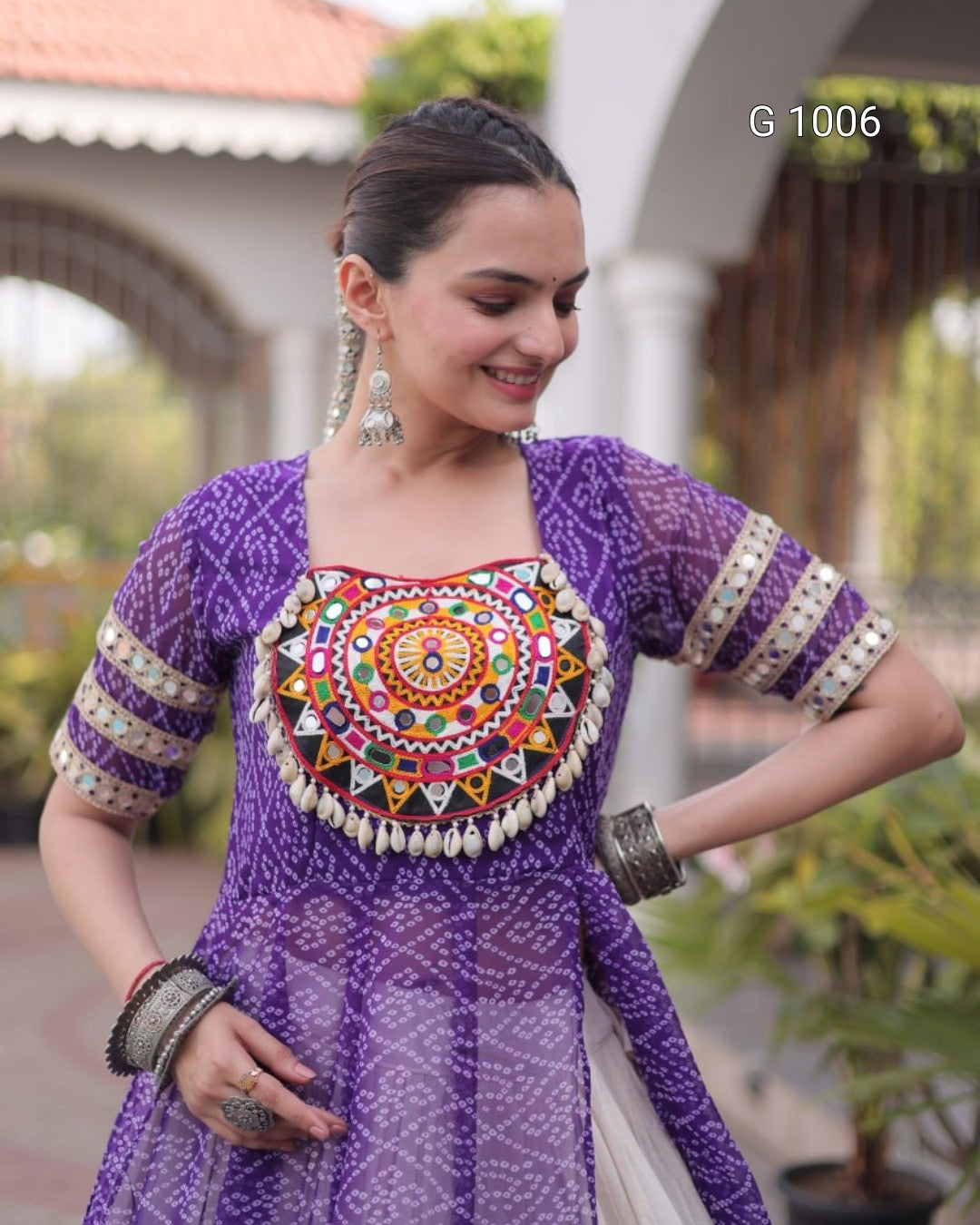 A woman wearing a purple embroidered traditional outfit with a heavily decorated multicolor chest panel, patterned sleeves, and silver jewelry, standing outdoors in front of a blurred architectural background.