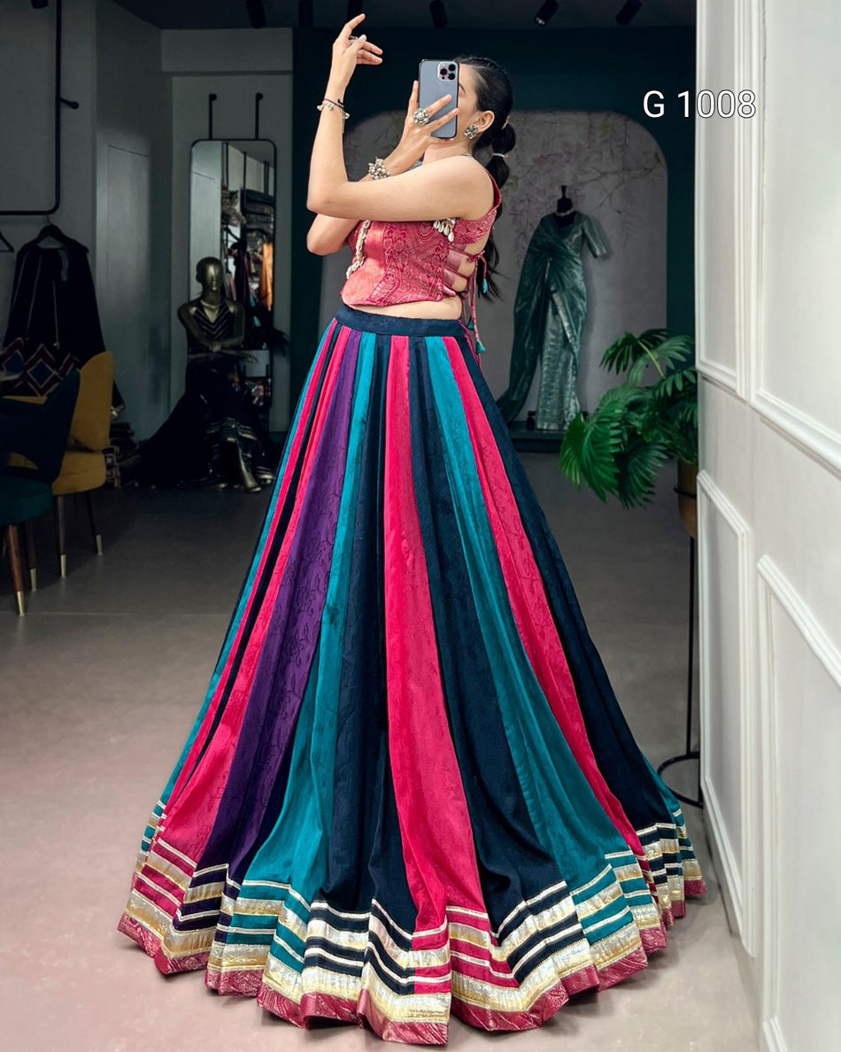 A woman takes a mirror selfie in a boutique wearing a vibrant, multi-colored traditional Indian skirt set (lehenga) with pink, teal, and navy blue vertical stripes and a matching embroidered crop top.