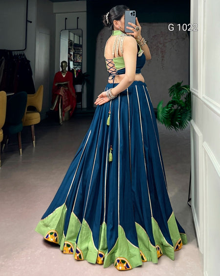 A woman takes a mirror selfie in a boutique wearing a traditional Indian lehenga choli set. The outfit features a navy blue flared skirt with green and gold patterned borders, a matching green lace-up choli, and gold jewelry.