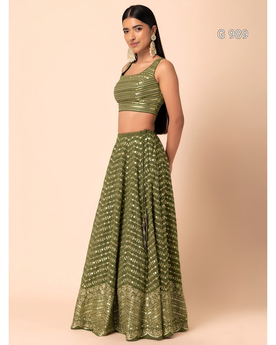A woman modeling a two-piece olive green sequined lehenga choli set against a solid beige studio background. The set features a sleeveless crop top and a long flared skirt with matching sequined patterns.