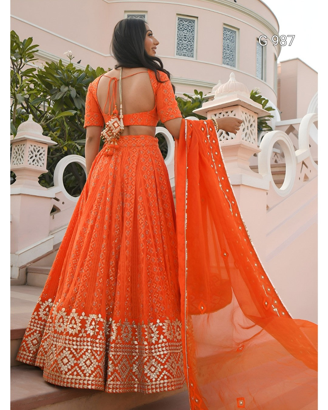 An orange embroidered Indian ethnic wear set consisting of a flared lehenga skirt, a short-sleeve crop blouse, and a matching dupatta, shown on a model from the back against a pink architectural background.