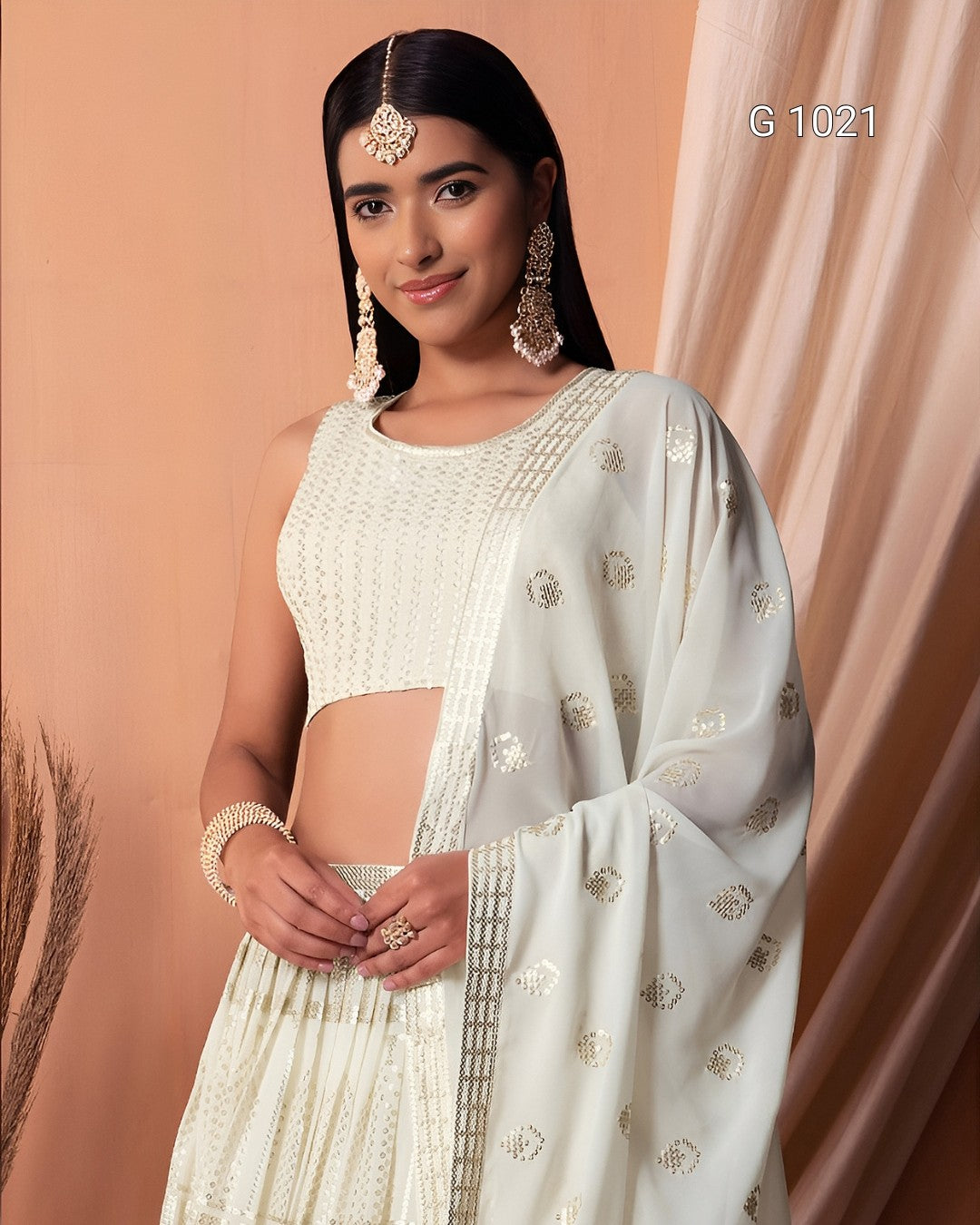 A woman modeling a traditional Indian outfit consisting of a white sleeveless embroidered blouse, a matching pleated skirt, and a sheer white dupatta with gold floral motifs, set against a peach-colored backdrop.