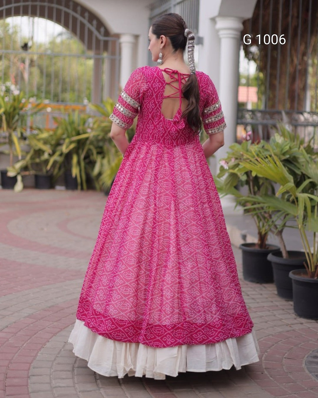 A woman wearing a bright pink traditional Indian dress with intricate embroidery and a white underskirt, photographed from the back in an outdoor courtyard setting.