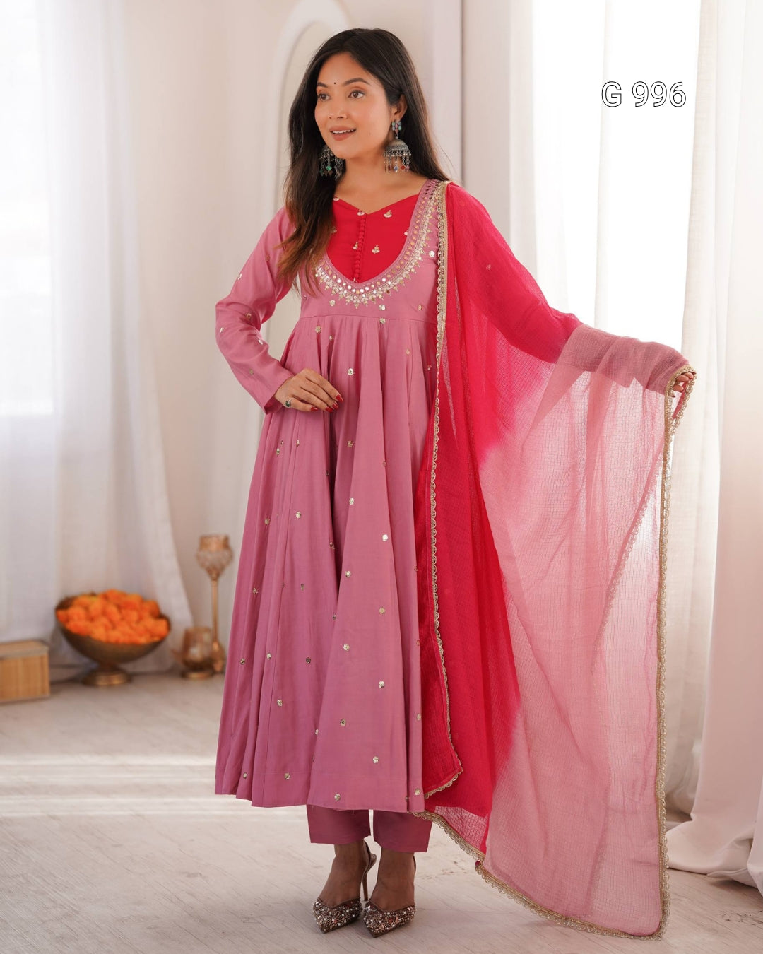 A woman models a traditional Indian outfit consisting of a dusty pink long kurta with silver embroidery, matching pink trousers, a bright pink inner lining, and a sheer pink dupatta with gold zari borders. She is standing in a brightly lit room with white curtains.