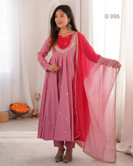 A woman models a traditional Indian outfit consisting of a dusty pink long kurta with silver embroidery, matching pink trousers, a bright pink inner lining, and a sheer pink dupatta with gold zari borders. She is standing in a brightly lit room with white curtains.