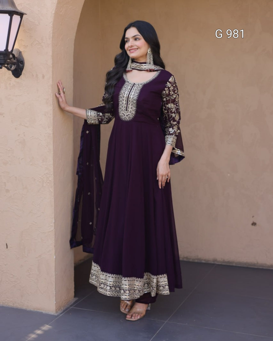 A woman models a deep purple Anarkali gown with ornate silver and gold embroidery, paired with a matching dupatta, set against a beige stucco wall background.