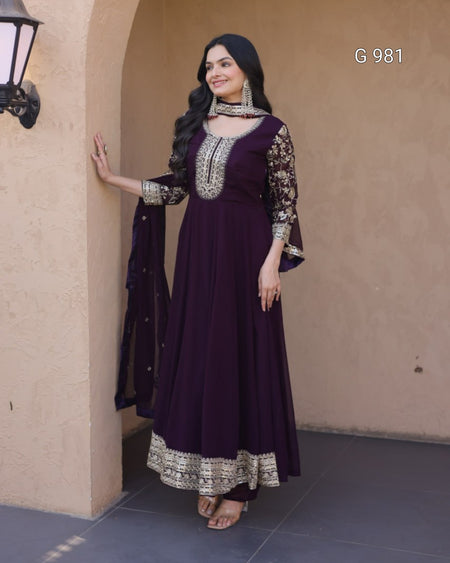 A woman models a deep purple Anarkali gown with ornate silver and gold embroidery, paired with a matching dupatta, set against a beige stucco wall background.